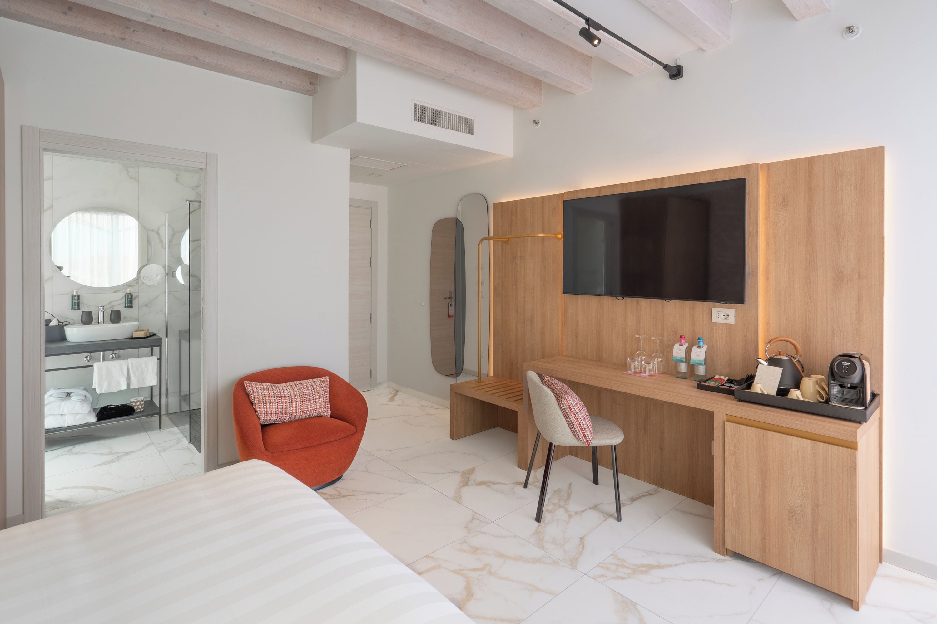 A modern bedroom with a bed, a desk with a chair, a TV, and a bathroom with a mirror and sink.