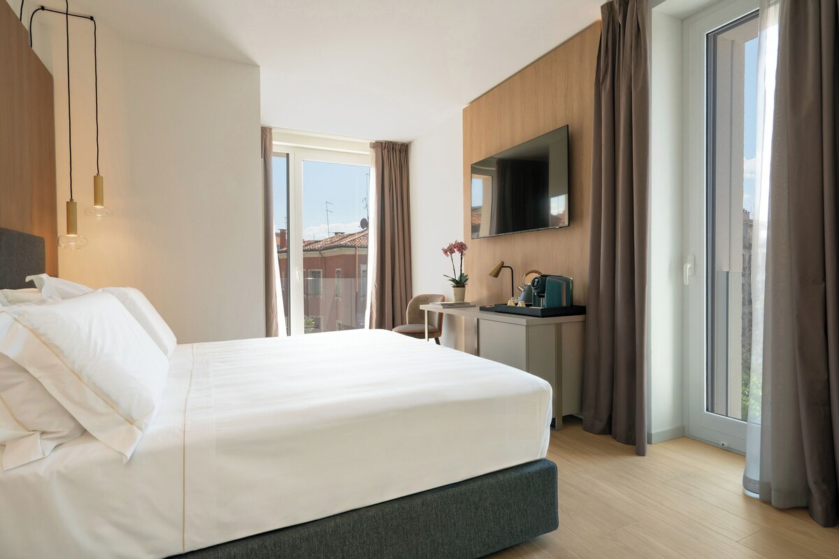 A modern hotel room with a large bed, a desk, a flat-screen TV, and a sliding glass door to the outside.