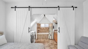 Quarto com cama, sofá cinza, máquina de lavar e banheiro com banheira e chuveiro.