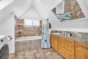 A bathroom with a shower, a bathtub, a washing machine, wooden cabinets, and a granite countertop. - The Beach House at Mission Bay (San Diego)