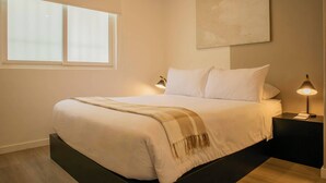 Ein ordentlich bezogenes Bett mit weißer Decke und kariertem Überwurf, begleitet von zwei Nachttischlampen auf schwarzen Nachttischen.