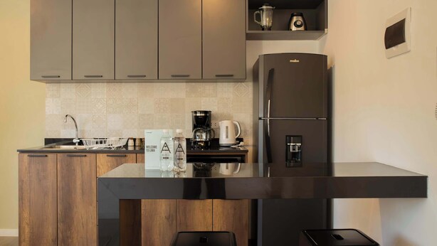 Cozinha moderna com ilha central, armários de madeira, geladeira preta, cafeteira e dois bancos pretos.