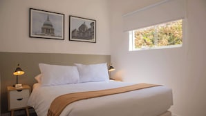 Ein Schlafzimmer mit einem großen Bett, zwei Nachttischen, einem Fenster mit Blick auf Bäume und zwei gerahmten Bildern an der Wand.