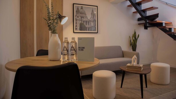 A modern living room with a round wooden table, a sofa, a small ottoman, a staircase, and a framed cityscape picture on the wall.