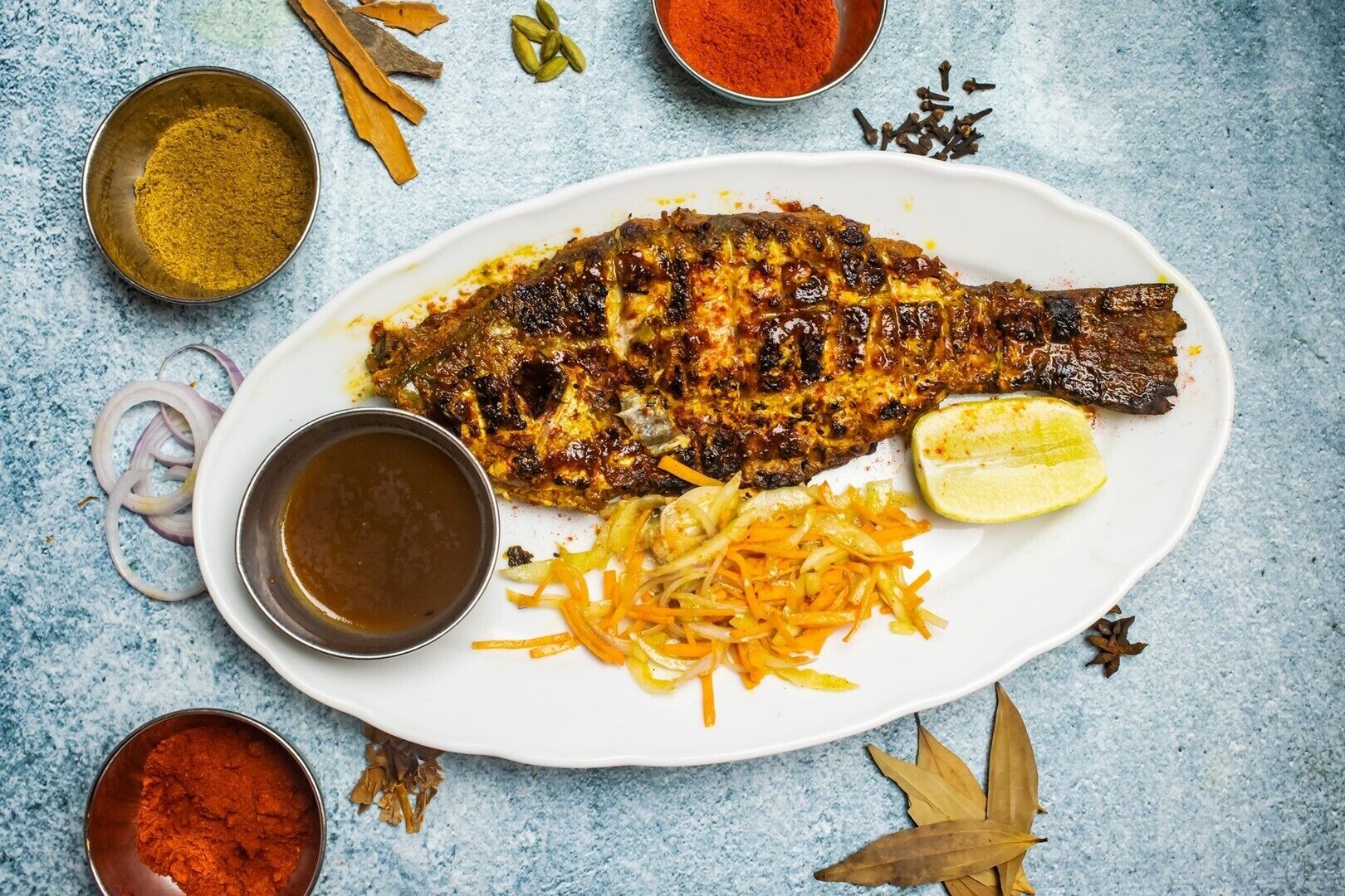 Prato de peixe grelhado com decoração de limão e cenoura, acompanhado por duas tigelas de molho, sobre uma superfície azul texturizada.