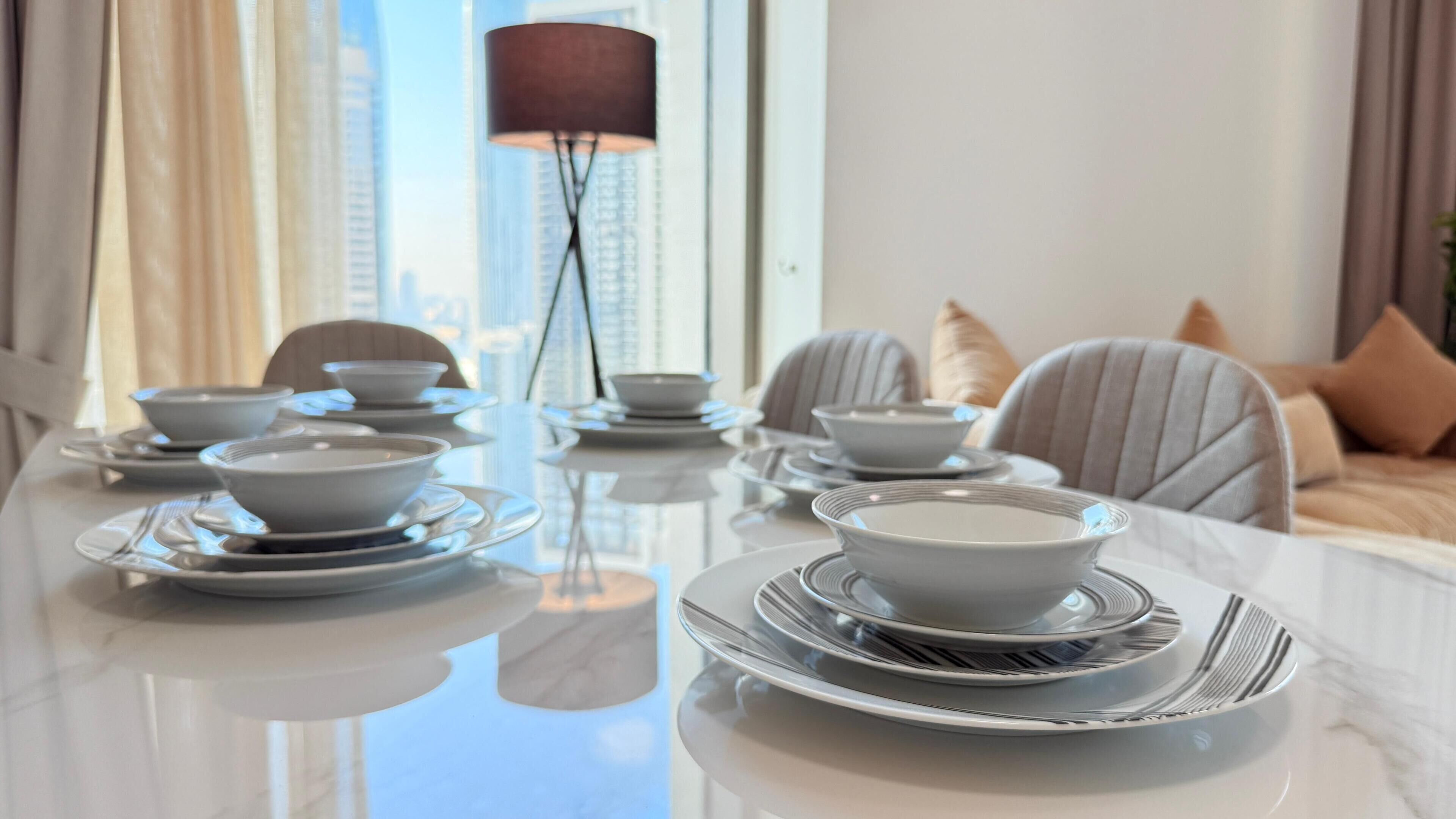 Mesa de comedor preparada con cuencos y platos blancos, una lámpara alta y vistas de la ciudad.