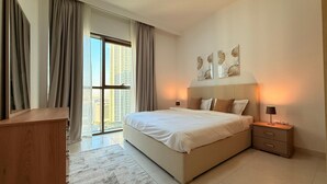 A modern bedroom with a large bed, a desk, and a view of tall buildings through the window. - Dancenter Luxury 2 BR at Creek Side (Dubai)