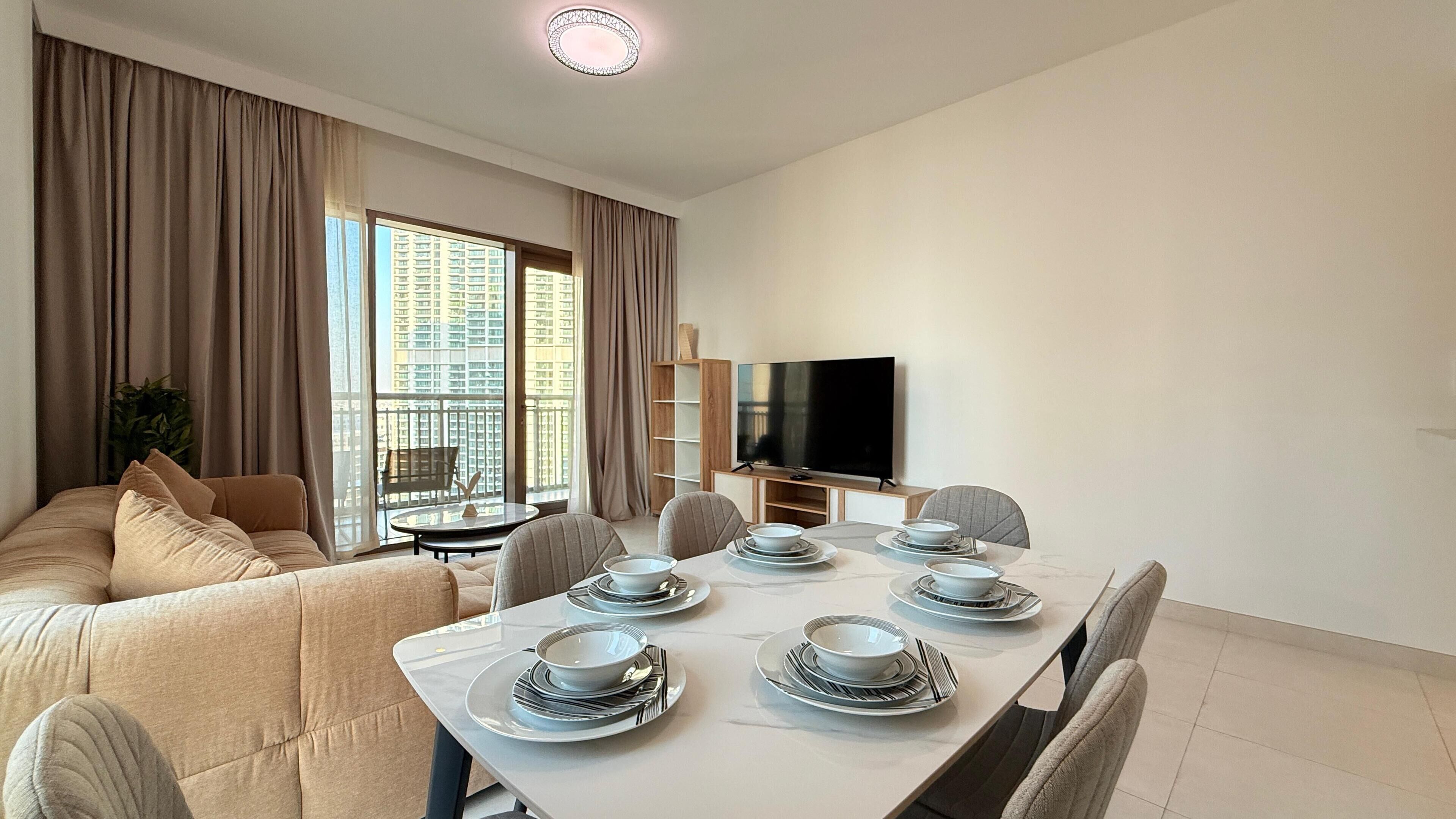 A modern living room with a sofa, dining table set with plates and bowls, a TV, and large windows with curtains.