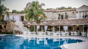 A swimming pool with lounge chairs and umbrellas, surrounded by stone buildings with large windows and a staircase leading to an upper level. - Nicolaus Prime Villas   (Castiadas)