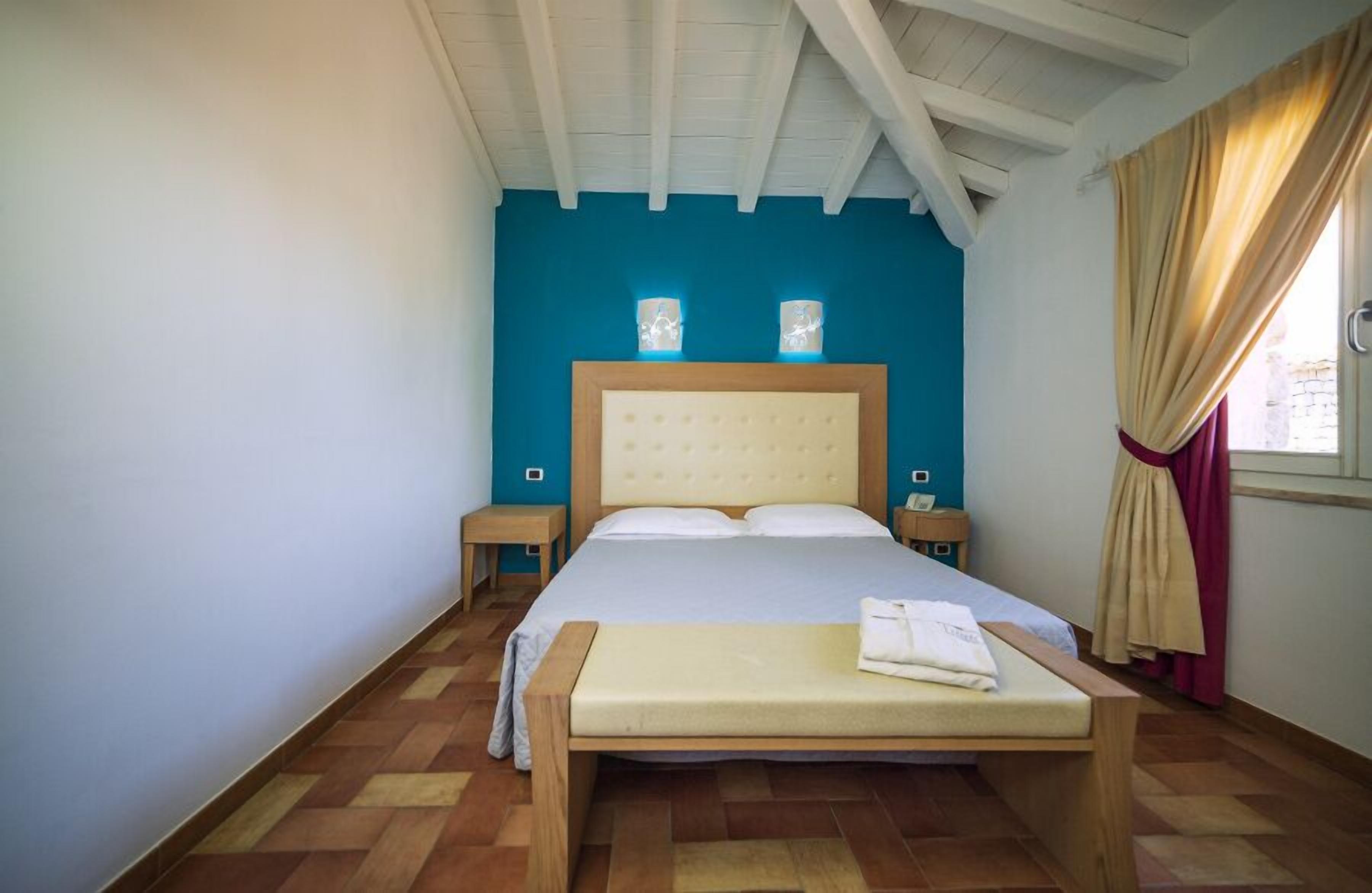 A bedroom with a large bed, two bedside tables, a bench, and a window with curtains.