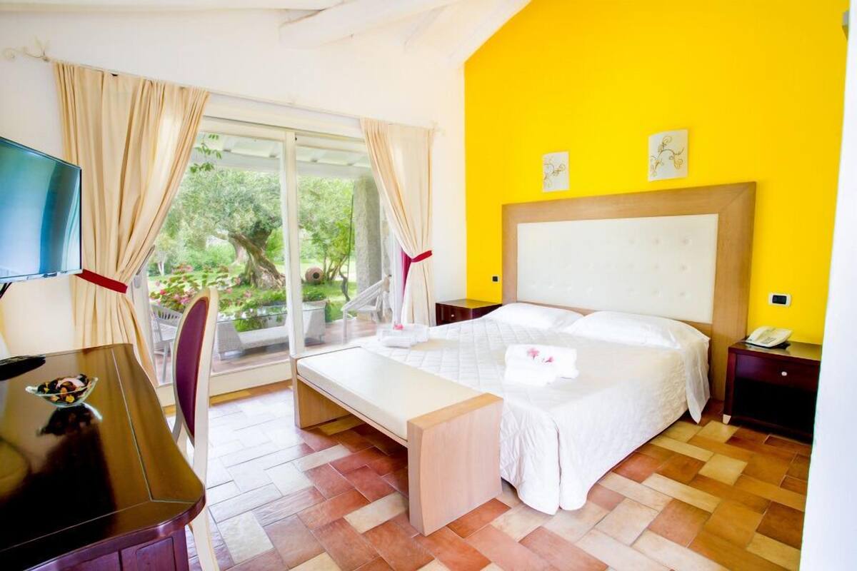 A hotel room with a bed, a desk, a TV, and a view of greenery outside.