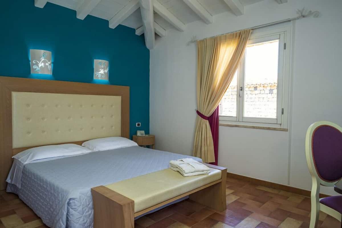 A bedroom with a bed, a nightstand, a bench, a chair, and a window with curtains.