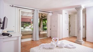A hotel room with a bed, a desk with a telephone, a large window with curtains, and a view of greenery outside. - Nicolaus Prime Villas   (Castiadas)