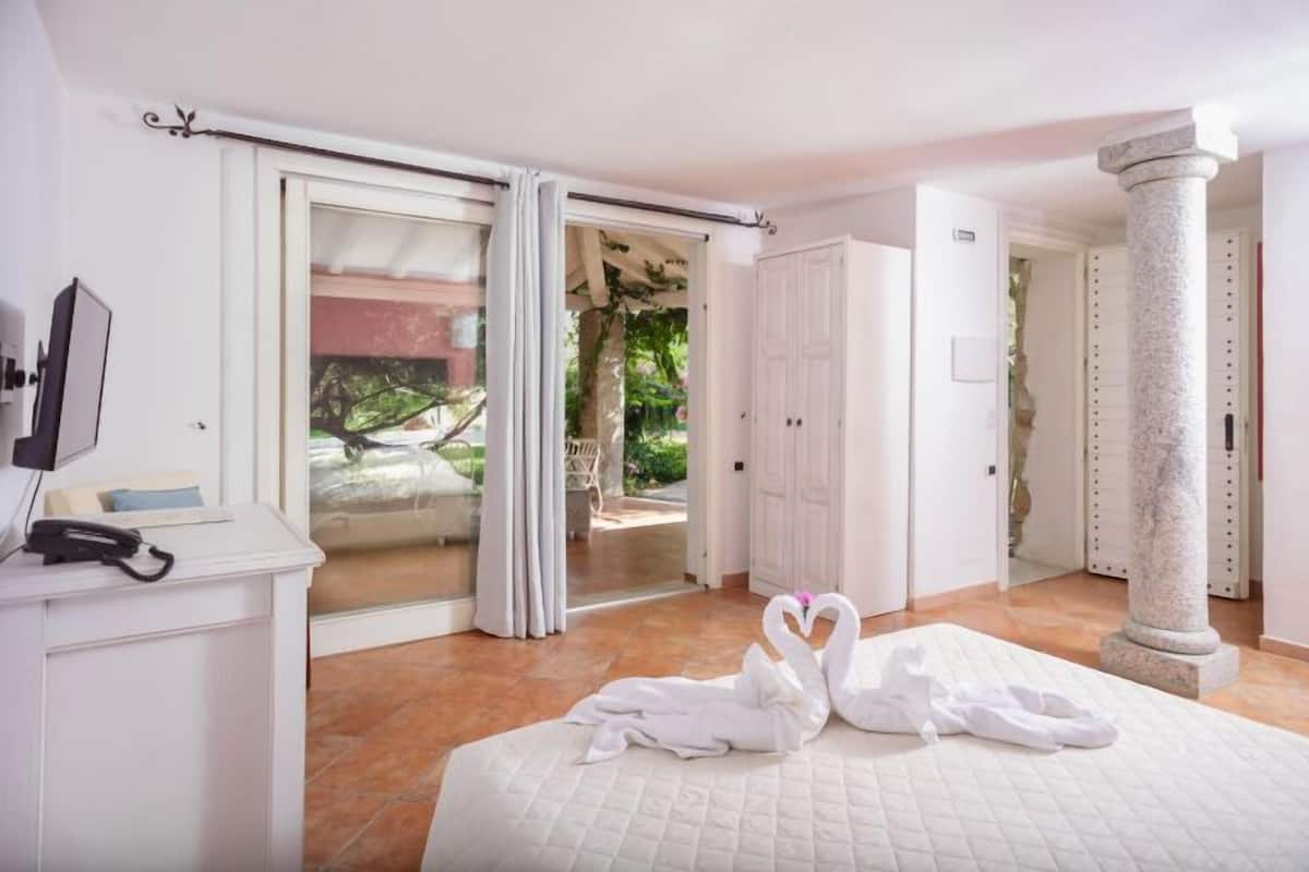 A hotel room with a bed, a desk with a telephone, a large window with curtains, and a view of greenery outside.