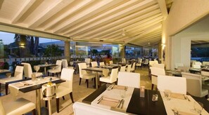 Een openluchtrestaurant met meerdere tafels en stoelen, grote ramen en een overdekt gedeelte.