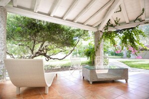 A covered patio with wicker furniture, a glass table, and a view of a garden with trees and pink flowers. - Nicolaus Prime Villas   (Castiadas)