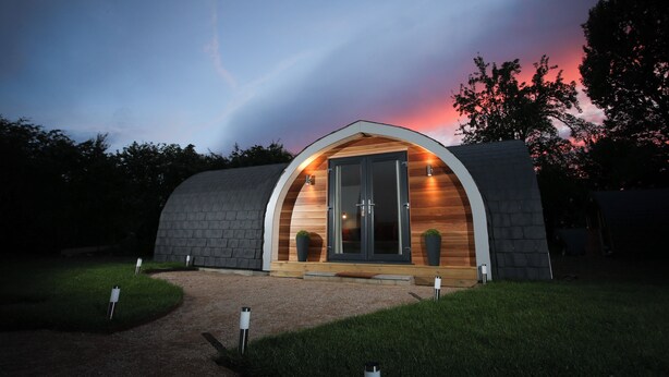 Un edifici modern, de forma semicircular, amb façana de fusta i una porta de vidre, envoltat d'un camí de grava i vegetació.