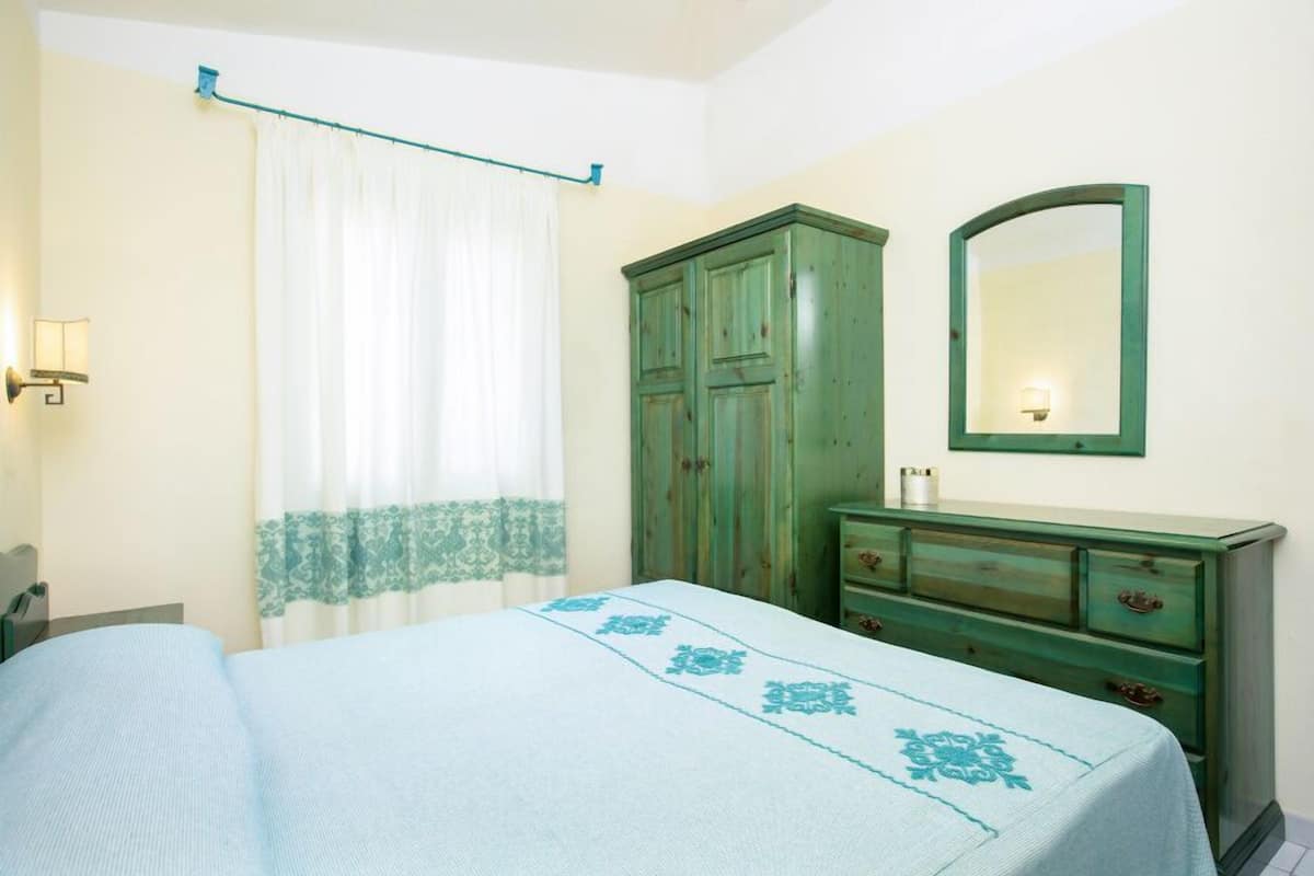 A bedroom with a bed, a green dresser, a mirror, and a window with curtains.