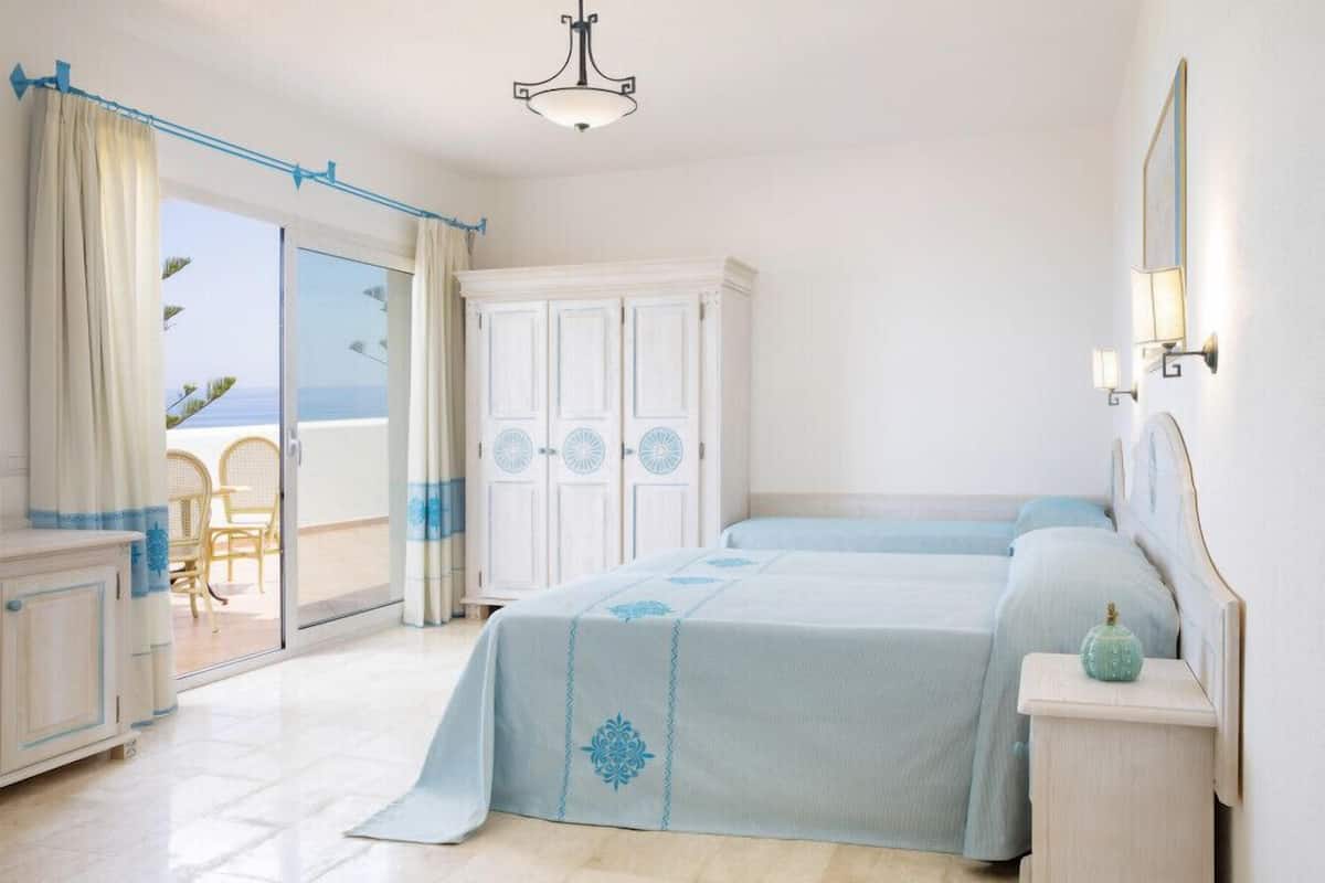 A bedroom with a balcony, a bed with a blue bedspread, a nightstand, and a wardrobe.