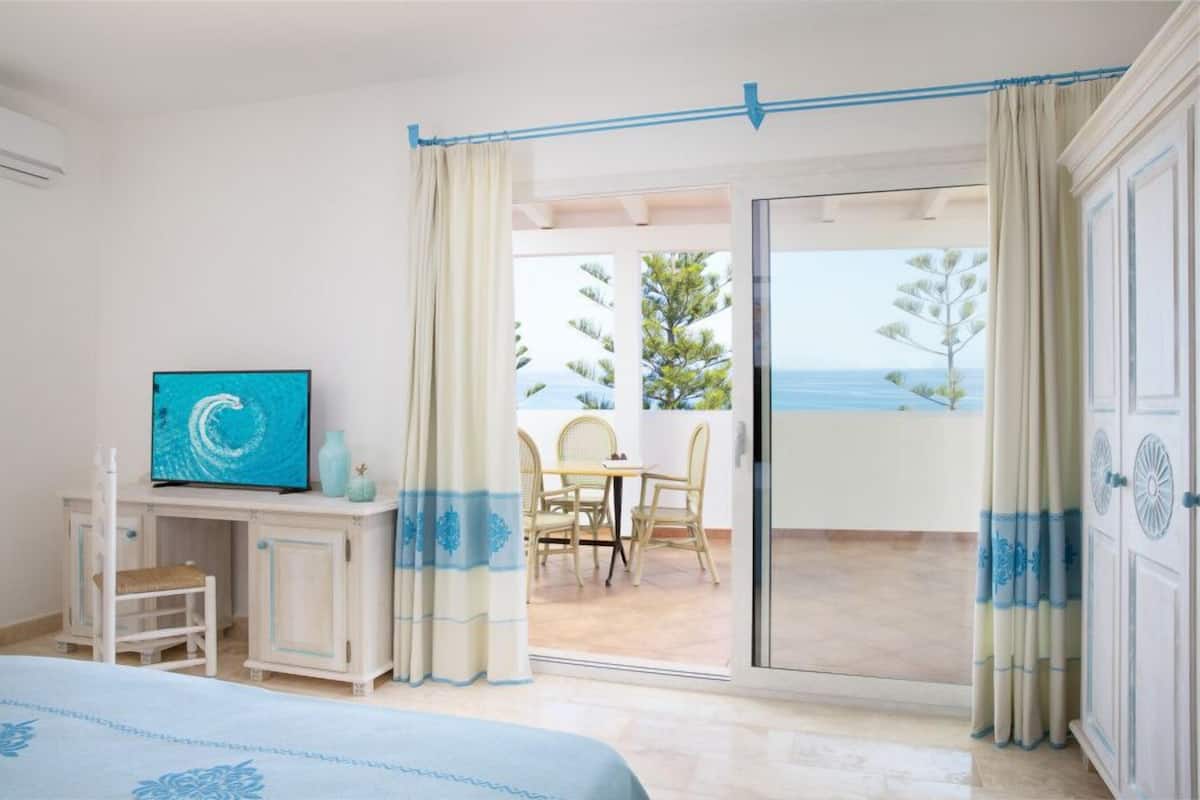 A hotel room with a bed, a desk with a TV, a balcony with a table and chairs, and a view of the sea.