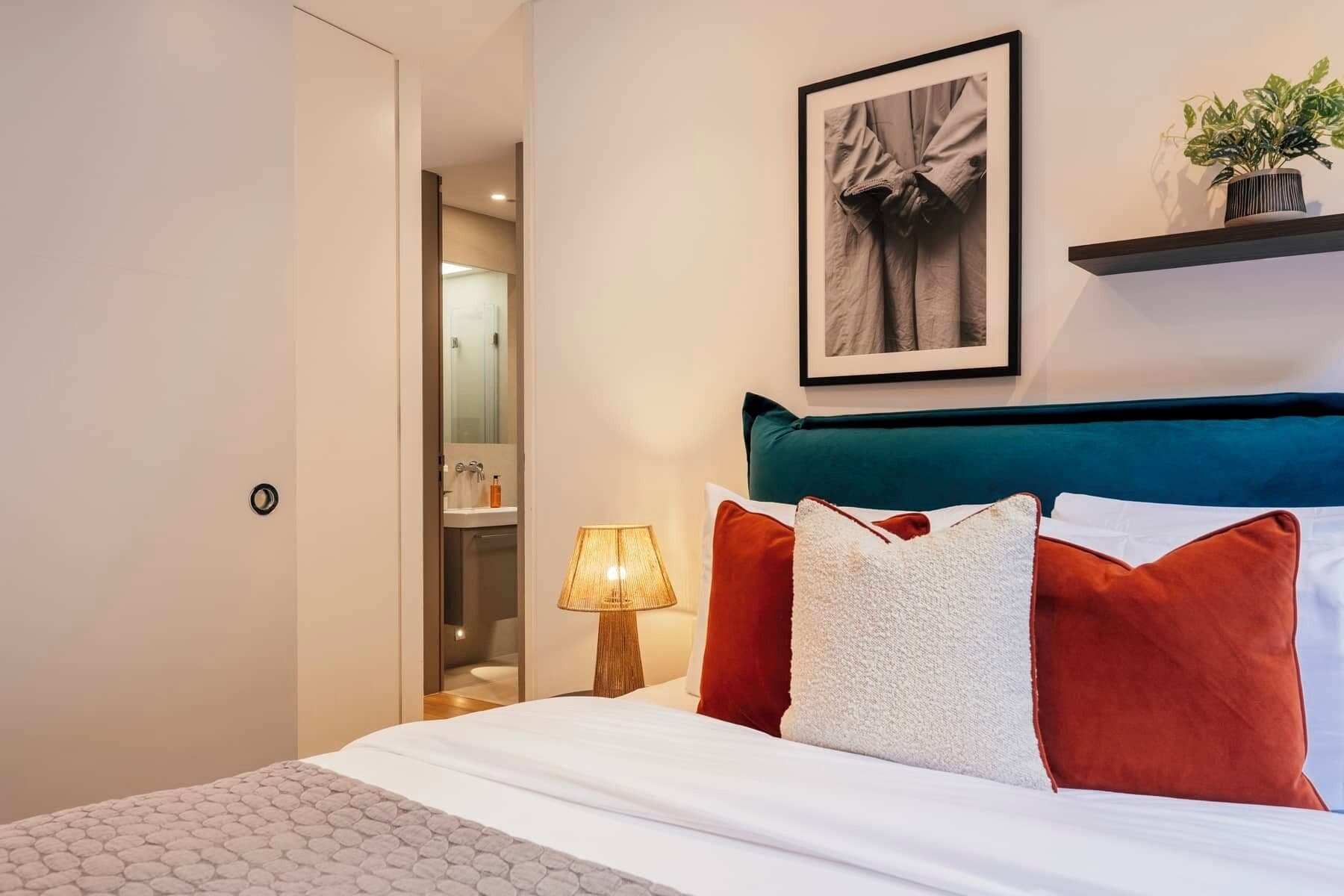 A hotel room with a bed, a nightstand, a lamp, a framed picture, and a plant on a shelf.