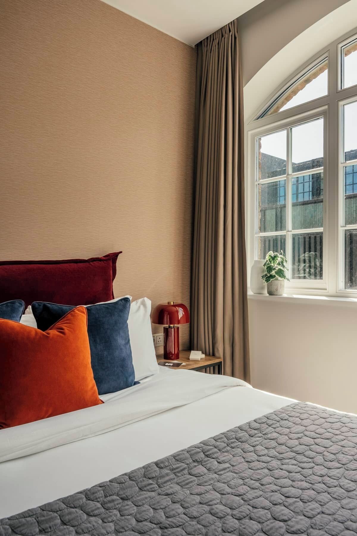 A hotel room with a bed, pillows, a lamp, a window with curtains, and a plant.