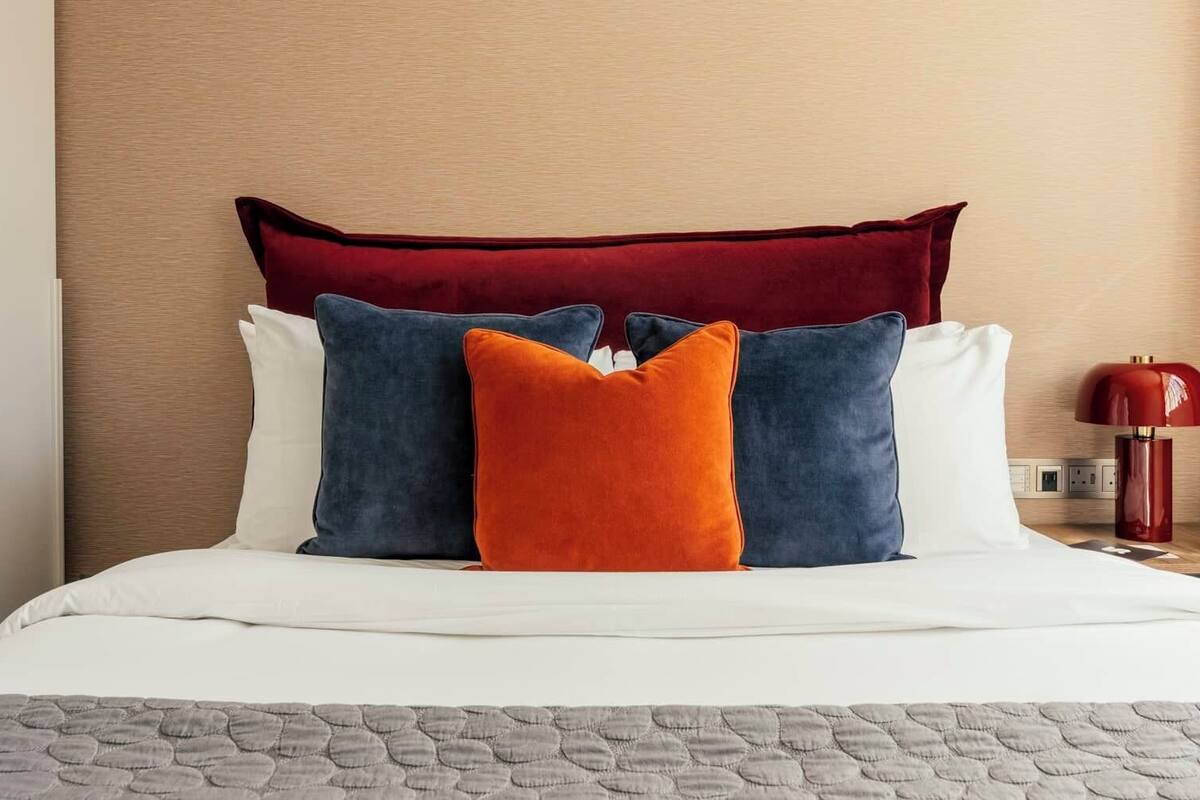 A neatly made bed with a white duvet, a grey quilt, and four pillows in red, blue, and orange.