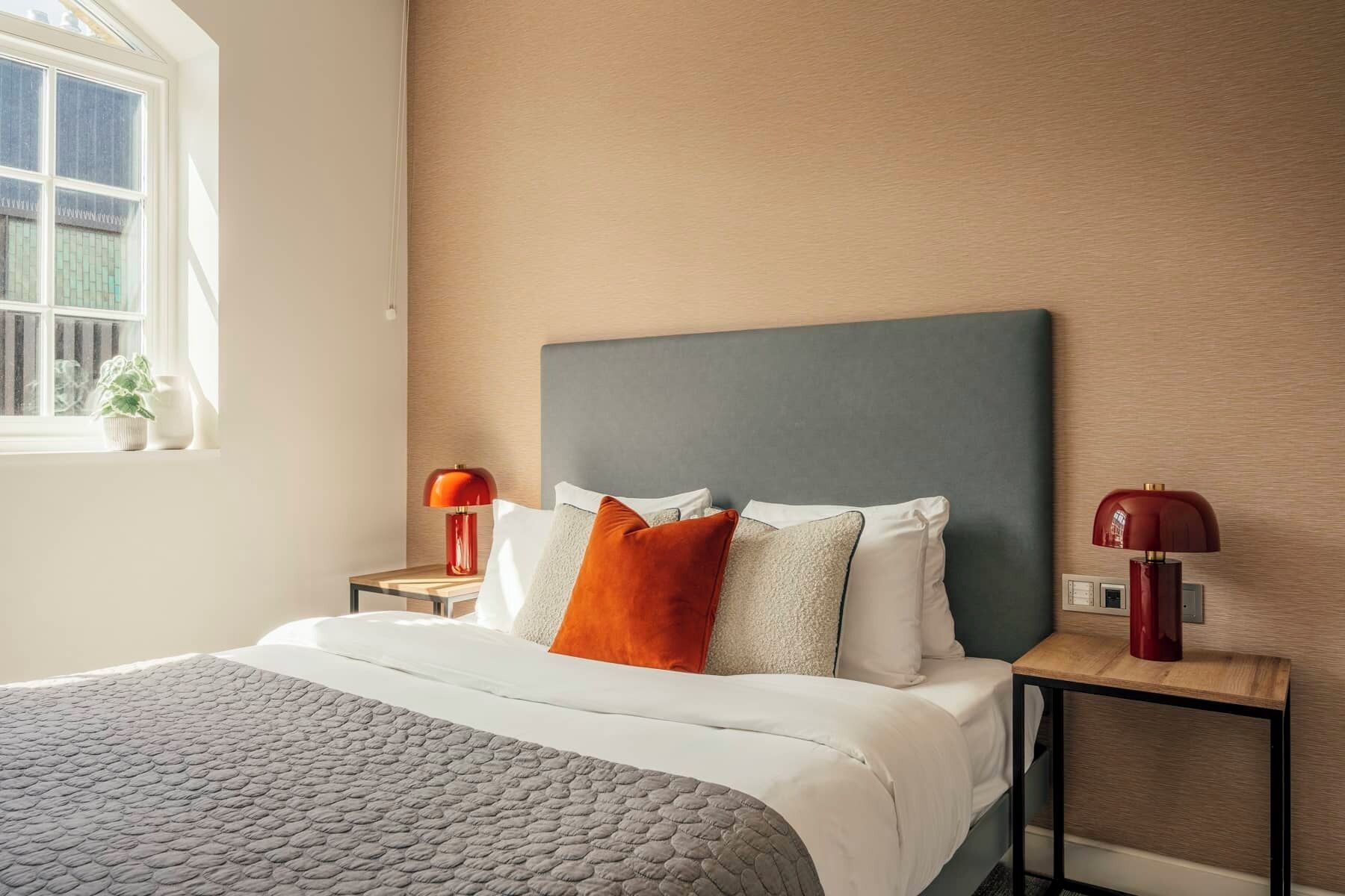 A neatly made bed with a grey quilt, a dark headboard, and two bedside tables with red lamps.