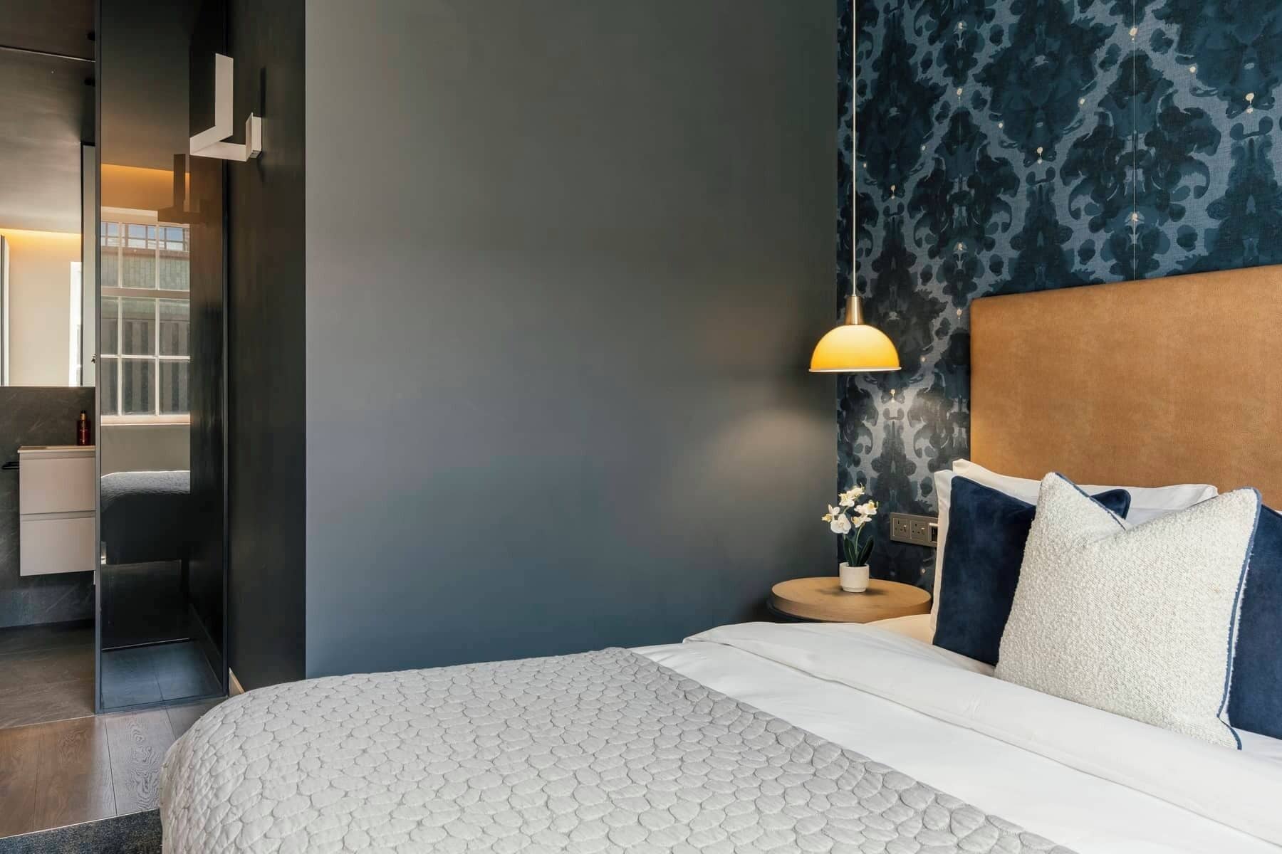 Una habitación de hotel moderna con una cama, una mesita de noche, una lámpara y un baño visible a través de una puerta abierta.