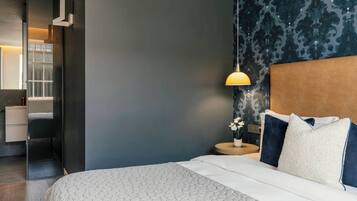 Una habitación de hotel moderna con una cama, una mesita de noche, una lámpara y un baño visible a través de una puerta abierta.