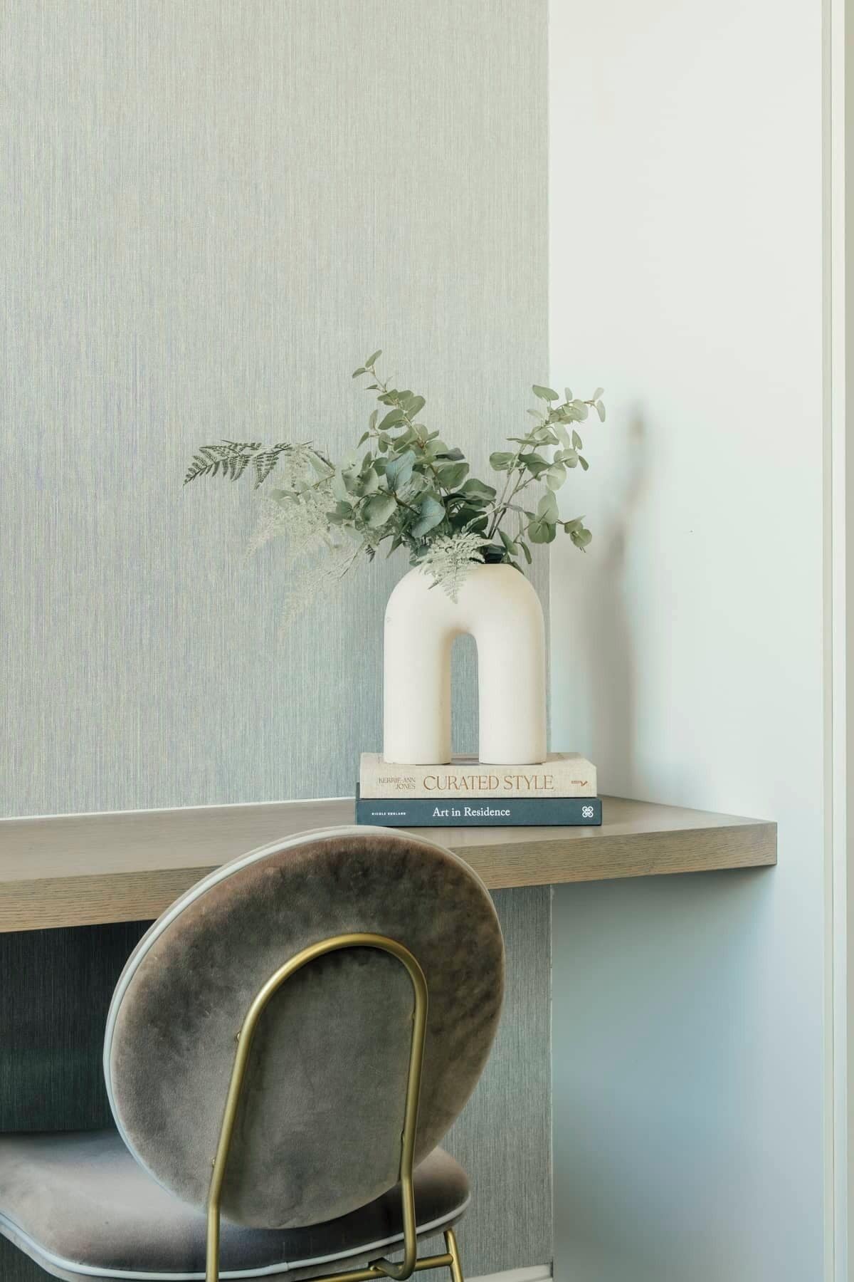 A bookshelf with books and a vase with green foliage, a chair with a gold frame, and a textured wall.