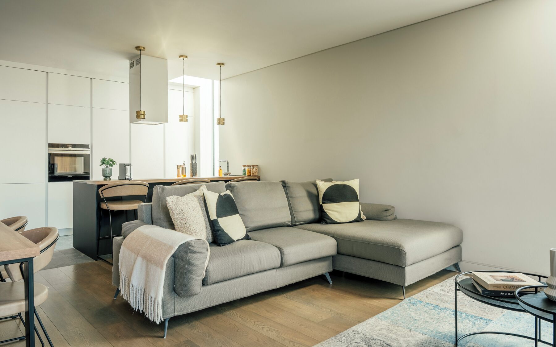 Una sala moderna con un sofá seccional gris, un pequeño comedor y una cocina con iluminación colgante.