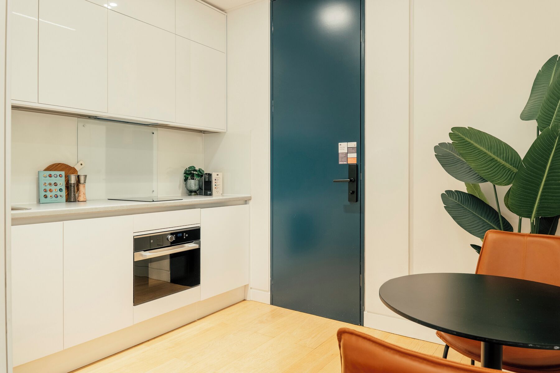 Una cocina moderna con armarios blancos, horno integrado, una puerta azul oscuro y una mesa redonda con una silla.