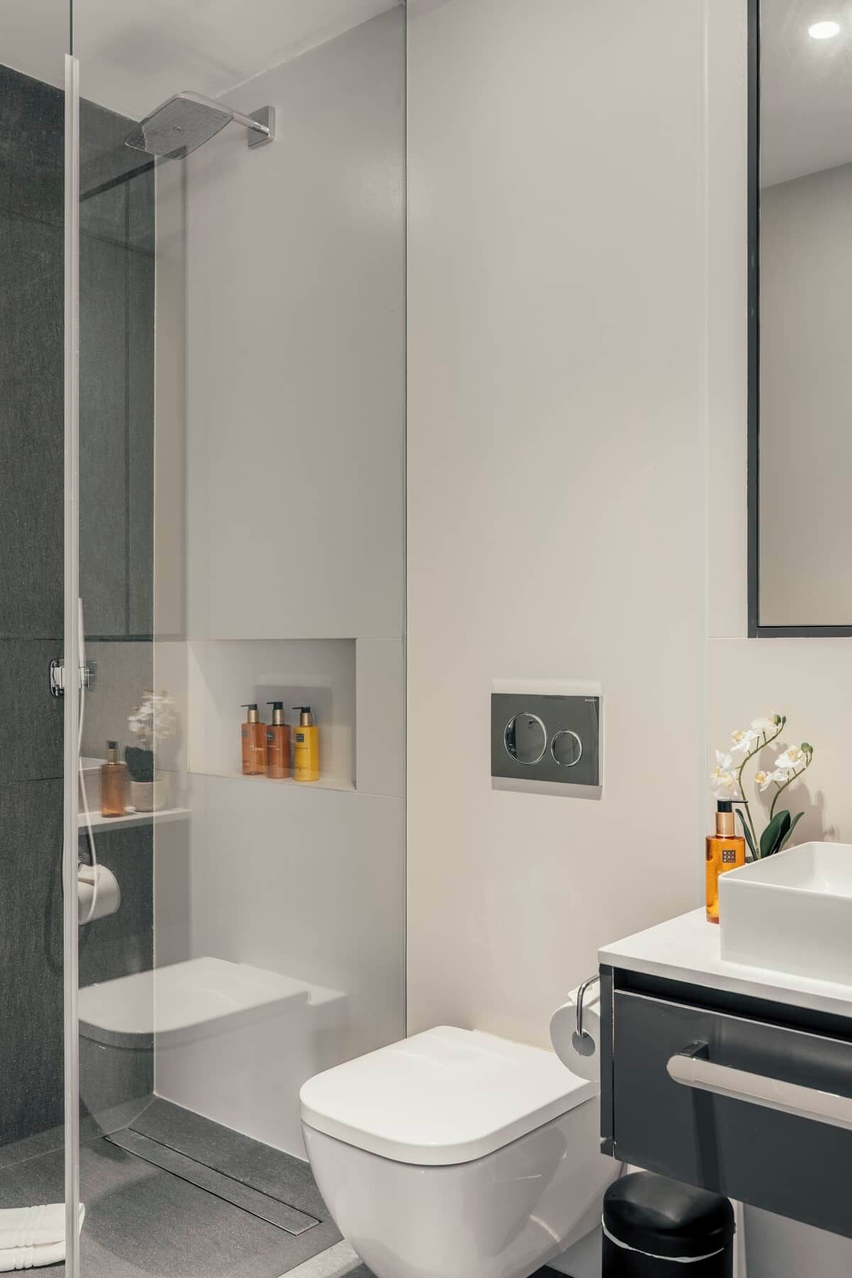 A modern bathroom with a glass shower enclosure, a toilet, a sink with a mirror, and a small shelf with toiletries.