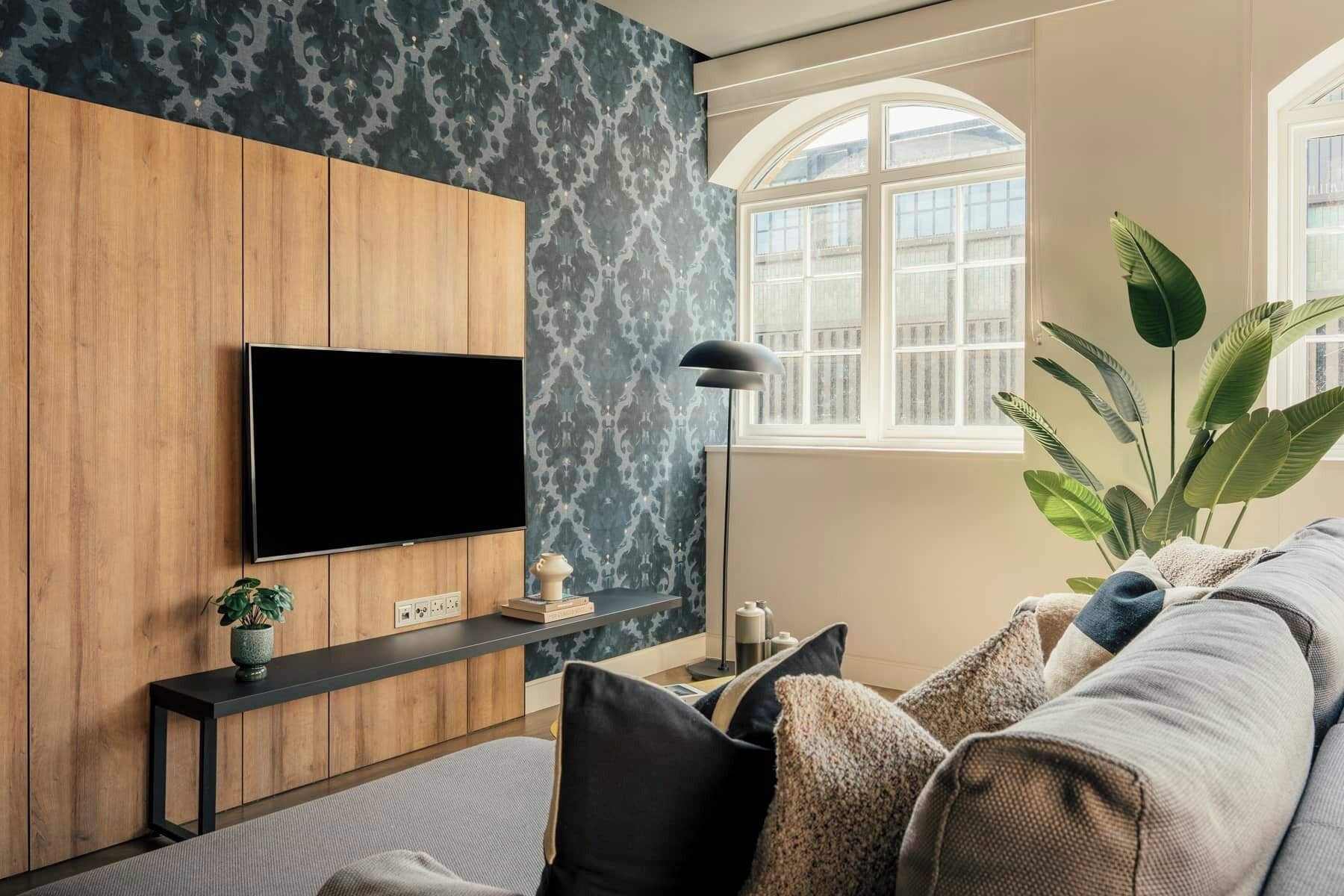 A living room with a TV mounted on a wooden panel wall, a floor lamp, a sofa with cushions, a potted plant, and a window with a view.