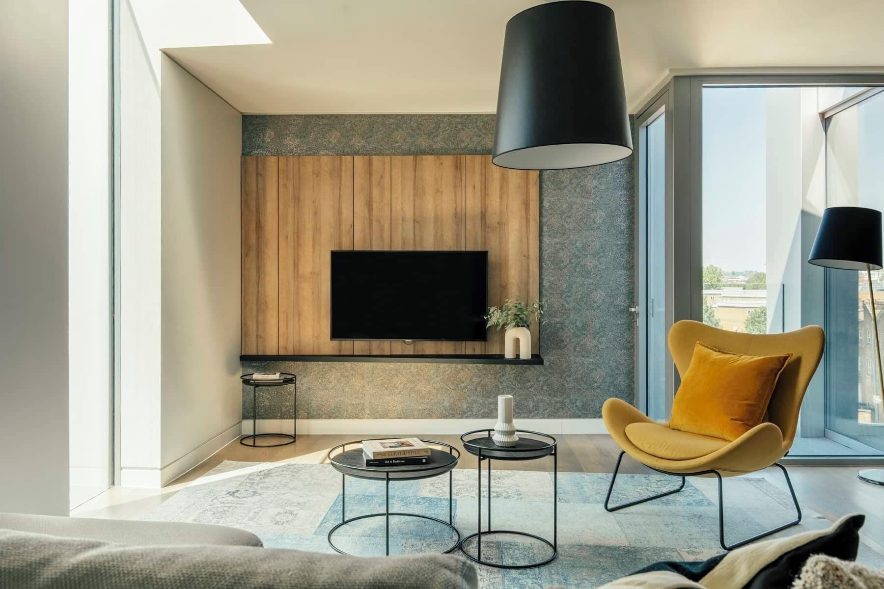 Een moderne woonkamer met een flatscreen televisie die aan een houten wand is bevestigd, een gele fauteuil, een ronde salontafel en een vloerlamp.
