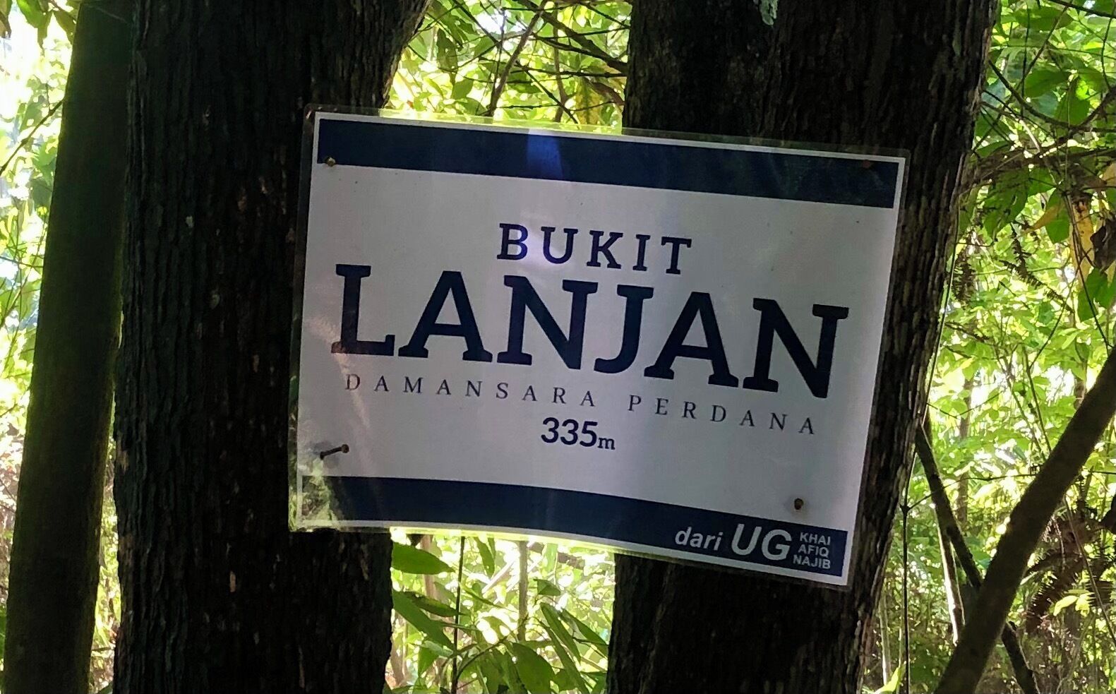 Señal que indica Bukit Lanjan, a 335 metros de Damansara Perdana, con árboles al fondo.