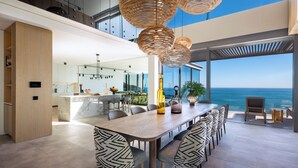 Een moderne eethoek met een lange houten tafel en gestreepte stoelen, een keuken op de achtergrond, en uitzicht op de oceaan door grote ramen.