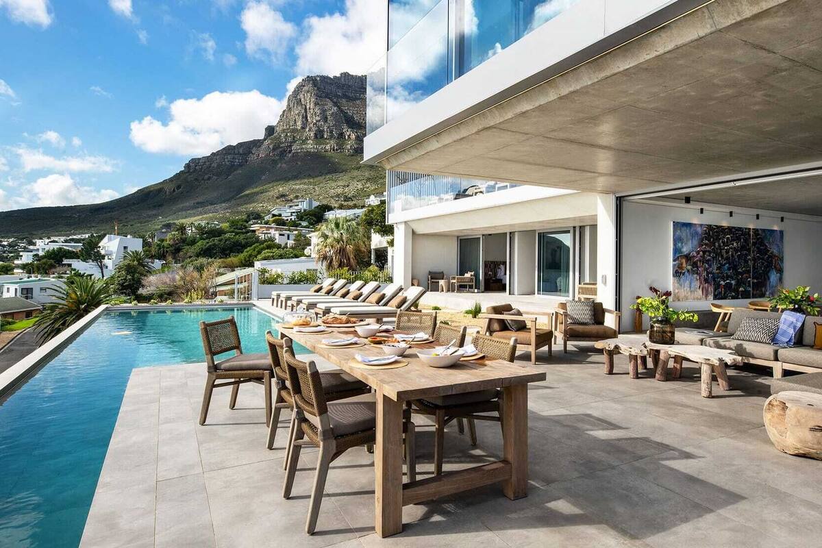 A modern outdoor dining area with a pool, lounge chairs, and a mountain view.