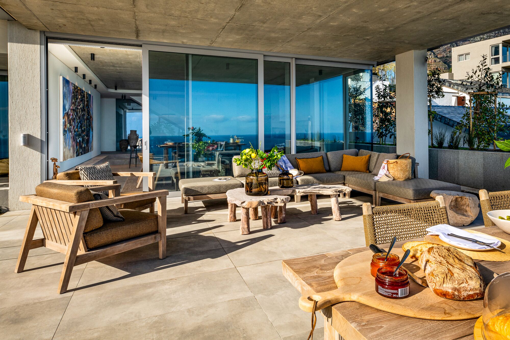 A modern outdoor seating area with a view of the ocean, furnished with wooden chairs and tables, and a dining setup with bread and jam.