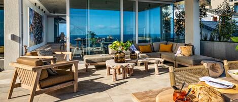A modern outdoor seating area with a view of the ocean, furnished with wooden chairs and tables, and a dining setup with bread and jam.
