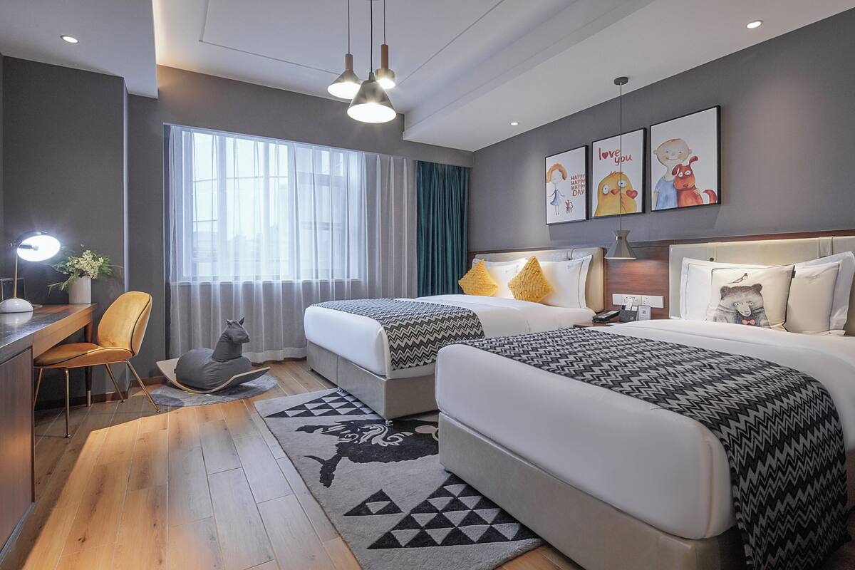 Una habitación de hotel moderna con dos camas, un escritorio con silla, un ventanal con cortinas y una obra de arte en la pared.