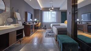 Ett modernt hotellrum med en stor säng, ett skrivbord med en stol, ett badrum med spegel och handfat samt ett stort fönster med gardiner.