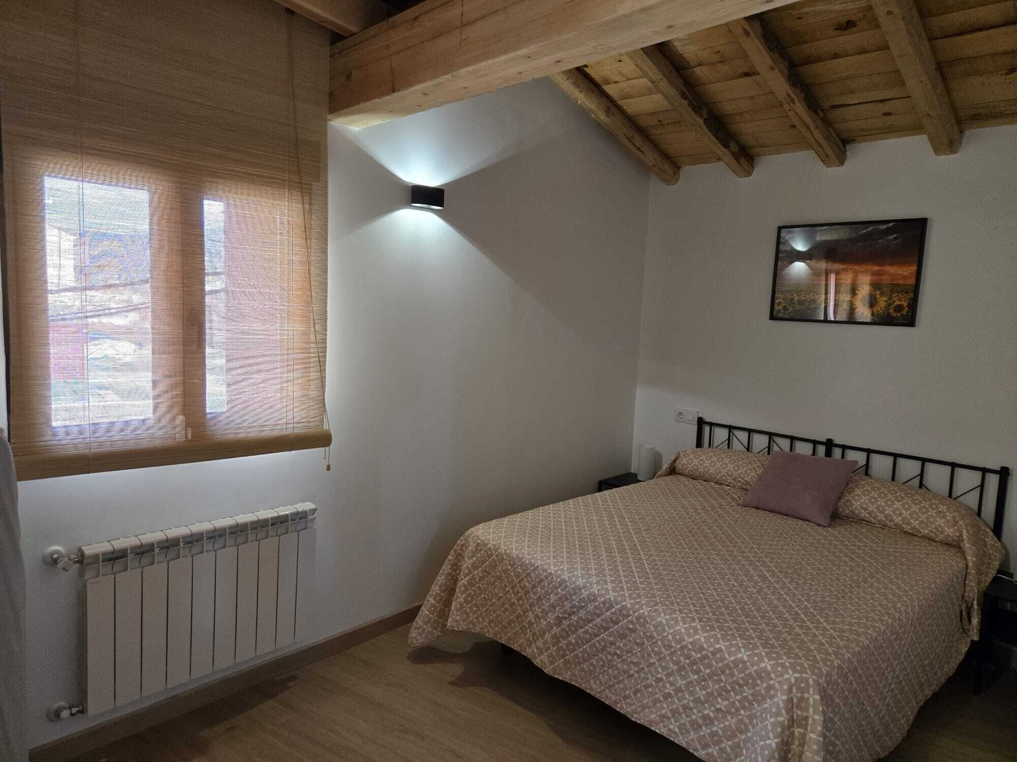 Un dormitorio con cama, radiador, ventana con persianas, luz de techo e imagen colgada en la pared.