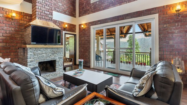 A spacious living room with a brick fireplace, leather sofas, a coffee table, and large windows offering a view of the outdoors. - Himvan by Akama Homes (Dhari)