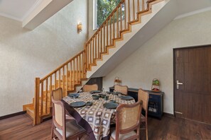A dining area with a wooden staircase, a dining table set with plates and bowls, and a cabinet with a basket on top. - Himvan by Akama Homes (Dhari)