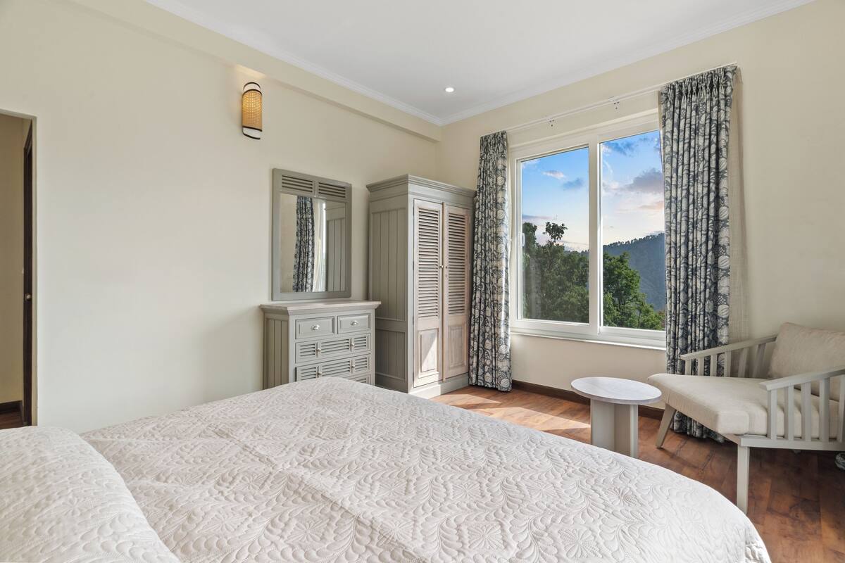 A bedroom with a large bed, a dresser, a chair, a small table, and a window with curtains.