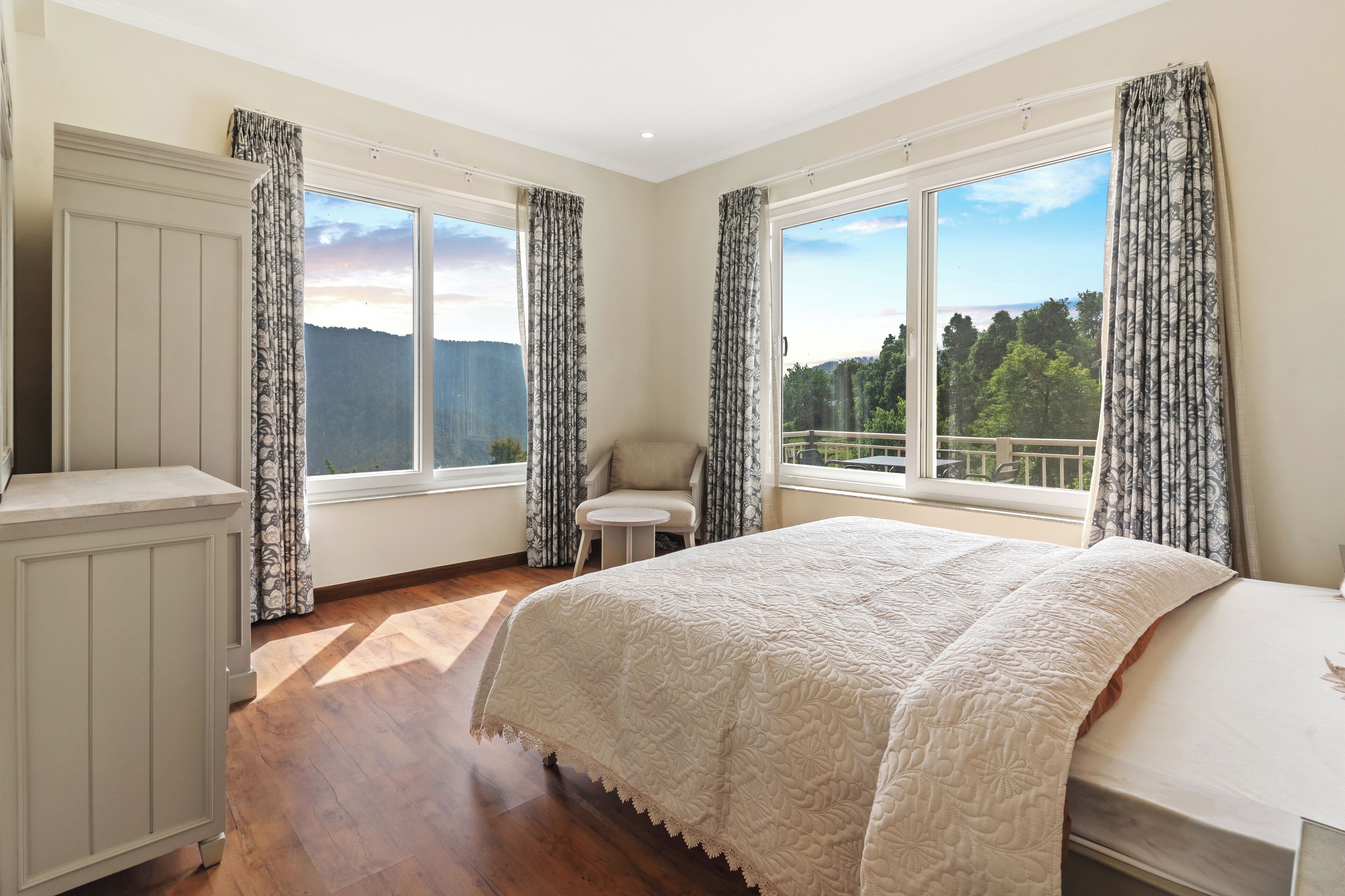 A bedroom with a large bed, a chair, and a view of the outdoors through large windows.