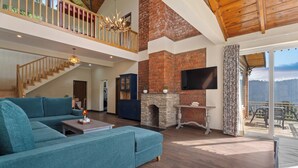 A spacious living room with a large teal sectional sofa, a stone fireplace, and a balcony with a view. - Himvan by Akama Homes (Dhari)