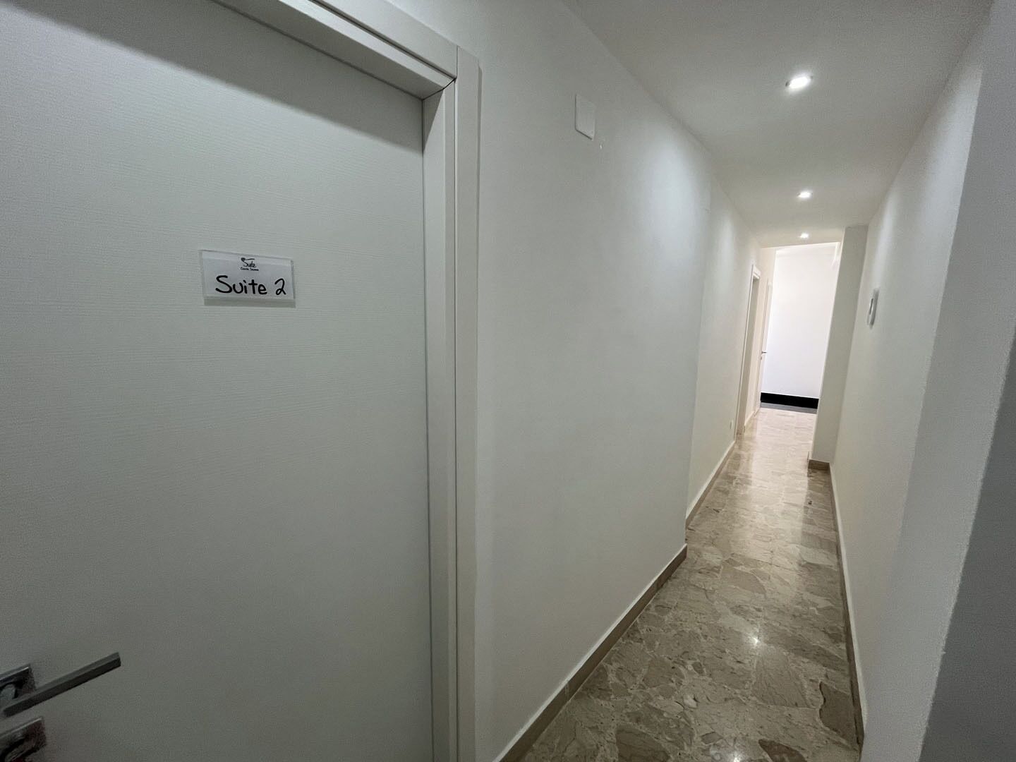 A hallway with a sign reading "Suite 2" on a white door. The walls are plain and the floor is tiled.